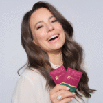 Zdjęcie, na którym ambasadorka produktu, Anna Mucha, trzyma w ręku bordową saszetkę kolagenu o smaku owoców leśnych. Modelka jest zadowolona, śmieje się.