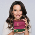 Zdjęcie, na którym ambasadorka produktu, Anna Mucha, trzyma w rękach pudełko zbiorcze kolagenu w proszku o smaku owoców leśnych. Modelka jest uśmiechnięta.