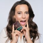 Zdjęcie, na którym ambasadorka produktu, Anna Mucha, trzyma w ręku brązową saszetkę kolagenu o smaku karmelowym. Modelka wydaje się pozytywnie zaskoczona.