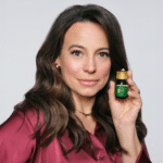 Ambasadorka produktu, Anna Mucha trzymająca w ręku produkt Glam4you shot o smaku brzoskwiniowym w szklanej butelce. Modelka patrzy wprost w obiektyw i jest delikatnie uśmiechnięta. Ma ciemne, falowane włosy, lekko za ramiona.