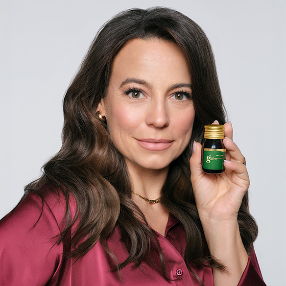 Ambasadorka produktu, Anna Mucha trzymająca w ręku produkt Glam4you shot o smaku brzoskwiniowym w szklanej butelce. Modelka patrzy wprost w obiektyw i jest delikatnie uśmiechnięta. Ma ciemne, falowane włosy, lekko za ramiona.