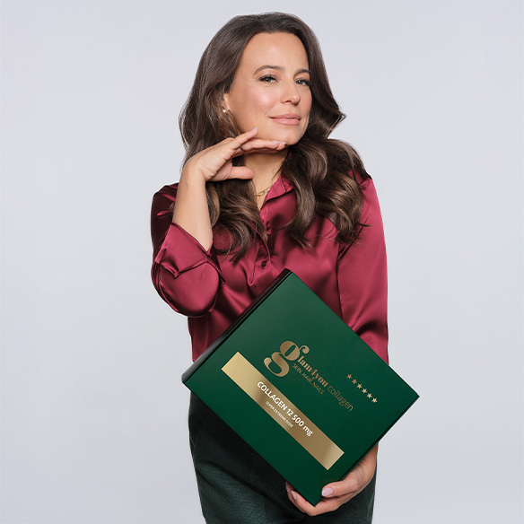 Ambasadorka produktu, aktorka Anna Mucha, trzymająca w jednej ręce zielone tekturowe pudełko produktu Glam4you Collagen shot o smaku brzoskwiniowym. Modelka drugą ręką podpiera w powietrzu podbródek.
